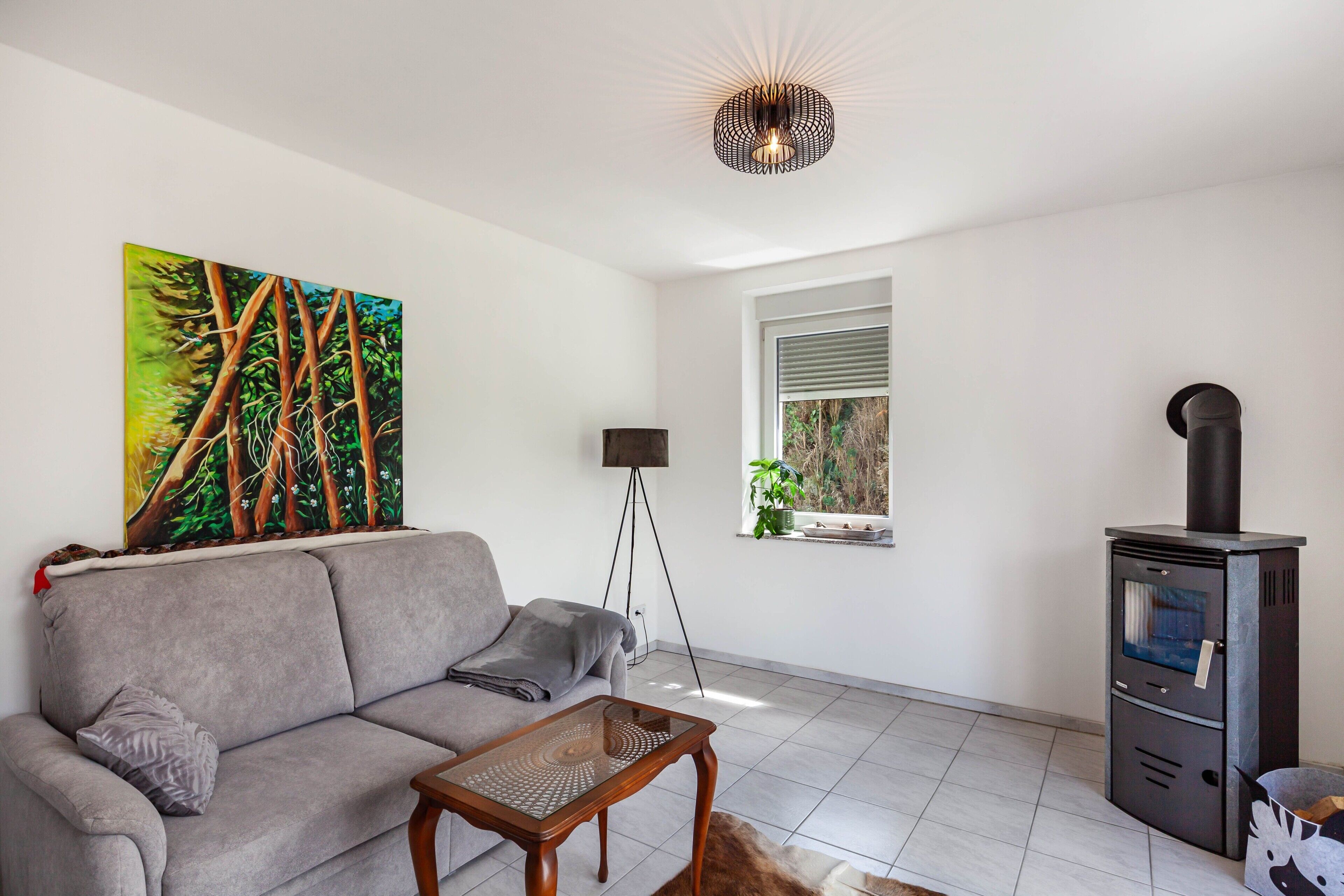 Un salon comprenant un canapé, une table basse, un tableau, une lampe sur pied, une fenêtre et un poêle à bois.