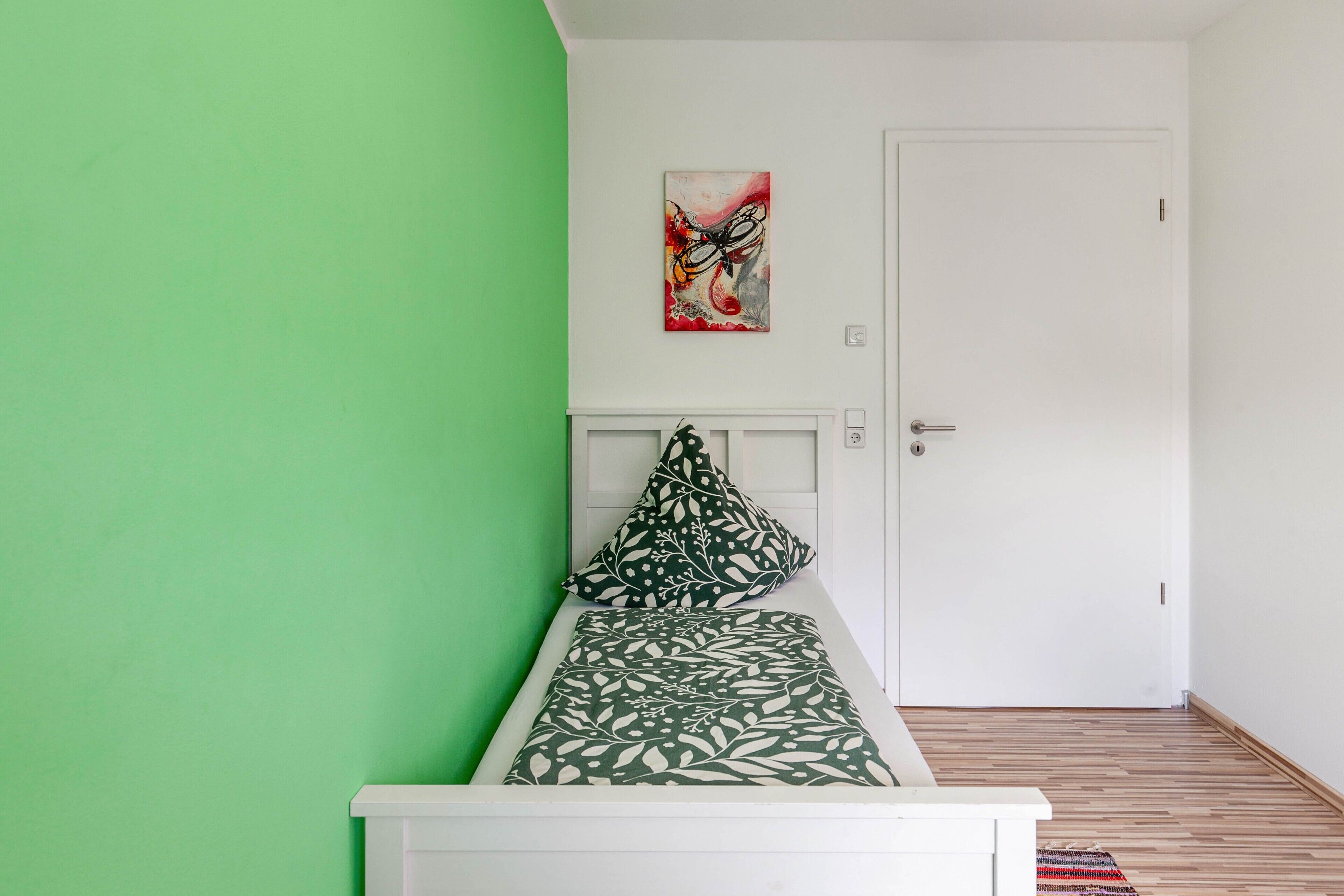 Un letto singolo con una coperta a motivi, un cuscino, un quadro appeso al muro e una porta bianca.