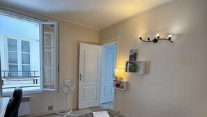 Ett sovrum med en säng, ett skrivbord, en stol, en bordsfläkt och ett fönster med utsikt över en balkong.