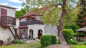 A two-story house with a balcony, a wooden staircase, and a garden with a tree and shrubs.