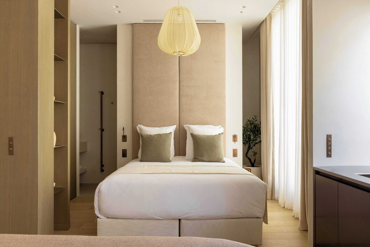 A modern bedroom with a large bed, a hanging light fixture, and a view of the bathroom.