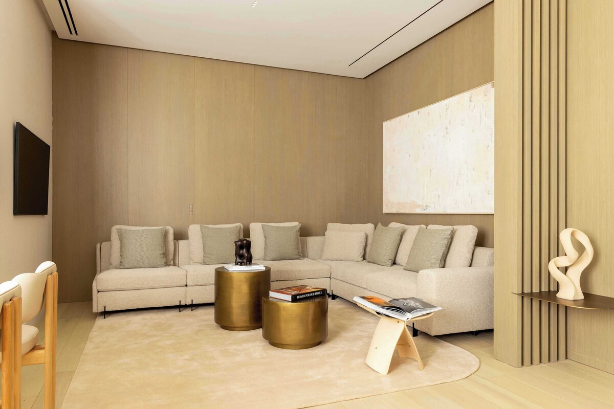 A modern living room with a sectional sofa, two gold side tables, a magazine on a wooden stand, and a minimalist sculpture.