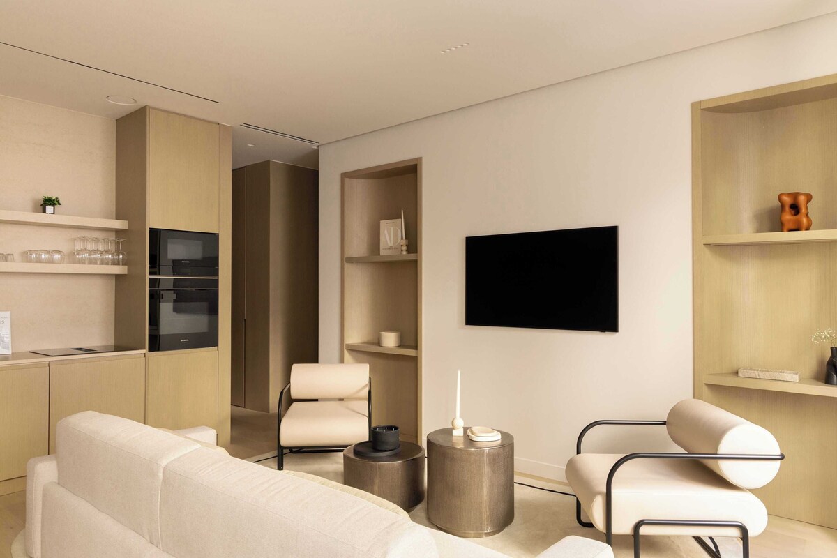 A modern living room with a sofa, two armchairs, a coffee table, a flat-screen TV mounted on the wall, and a kitchen area with a refrigerator and microwave.