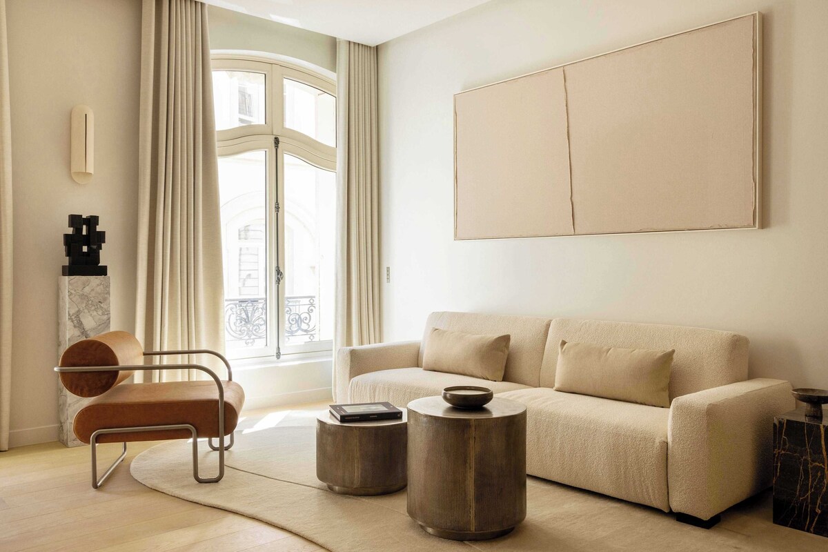 A modern living room with a sofa, two round side tables, a chair, and a large window with curtains.