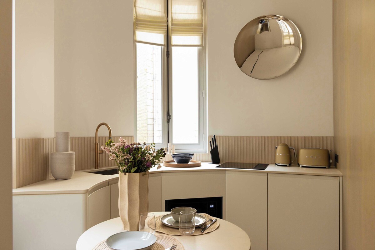 A modern kitchen with a dining area, a round mirror, and a window with blinds.