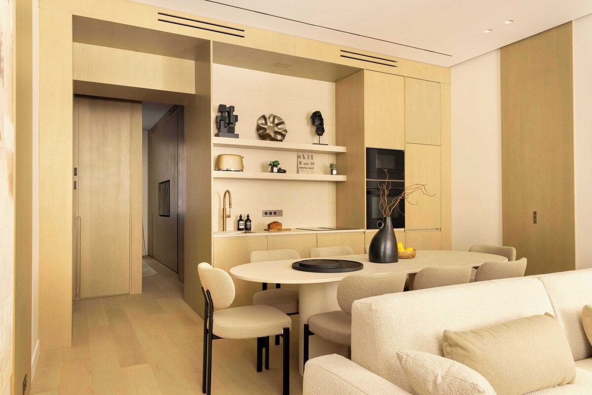 A modern living room with a sofa, a dining table, and chairs. There is a kitchen area with cabinets and a refrigerator.