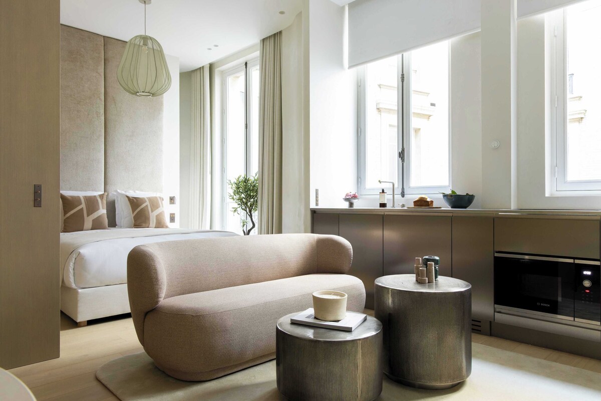 A modern hotel room with a large bed, a sofa, a dining area with a microwave, and large windows.
