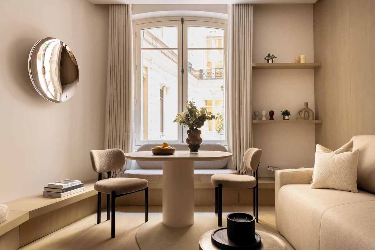A modern living room with a round table, two chairs, a sofa, a window with curtains, and a bookshelf with decorative items.