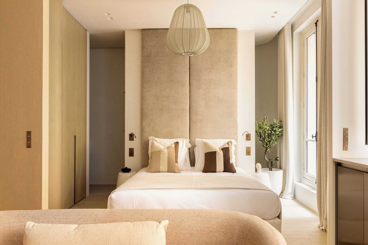 A modern hotel room with a large bed, a bedside table, a lamp, and a view of the outdoors.