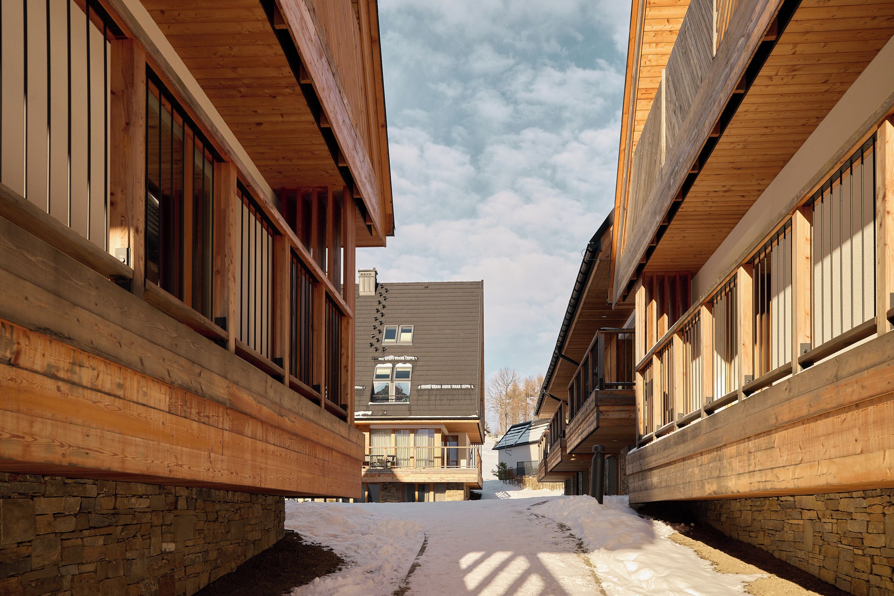 A narrow pathway between wooden buildings with stone walls, leading to a central building with a grey roof.