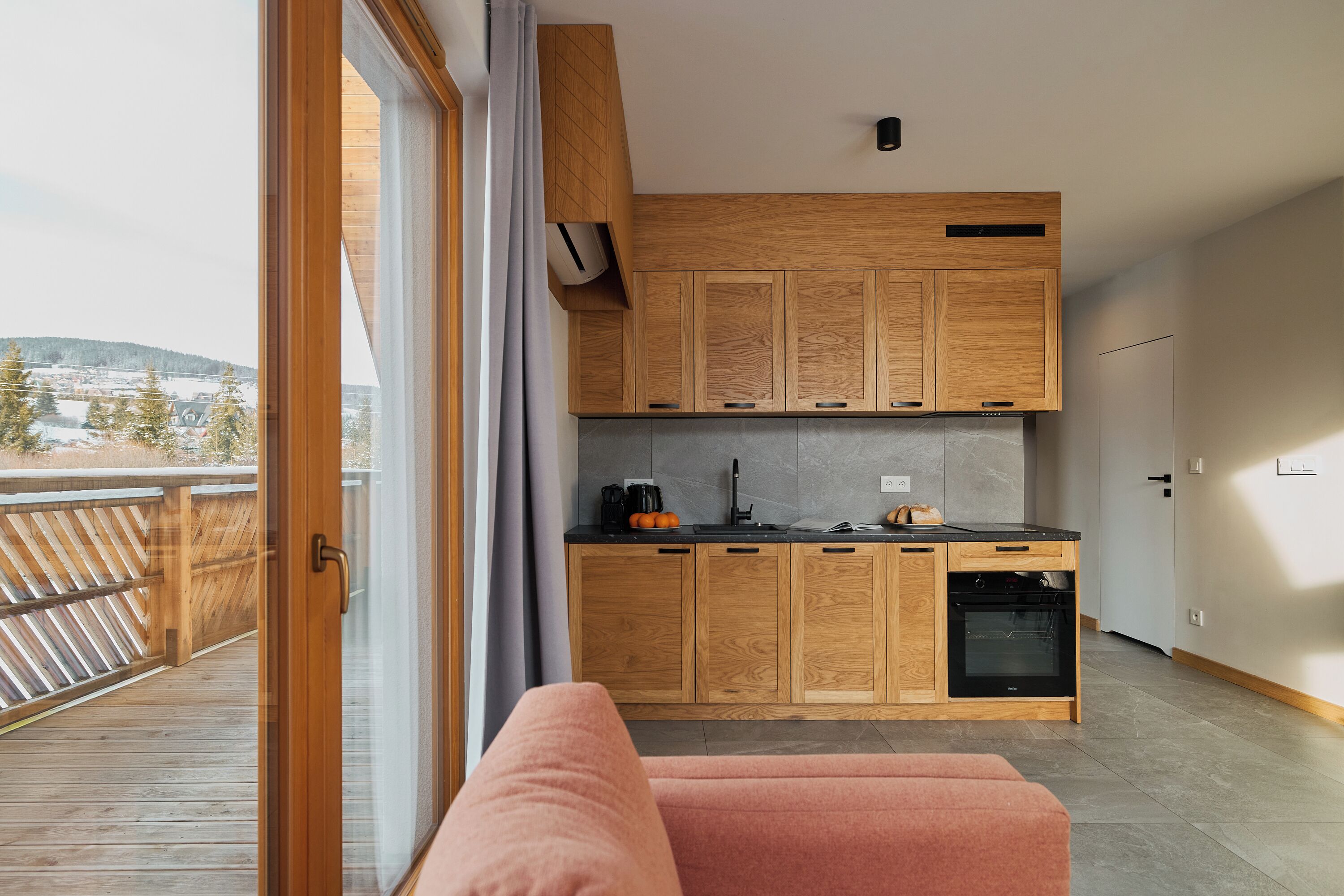 Eine moderne Küche mit Holzschränken und einem eingebauten Ofen, ein gemütliches rosa Sofa und Blick auf eine verschneite Landschaft durch eine Glastür.