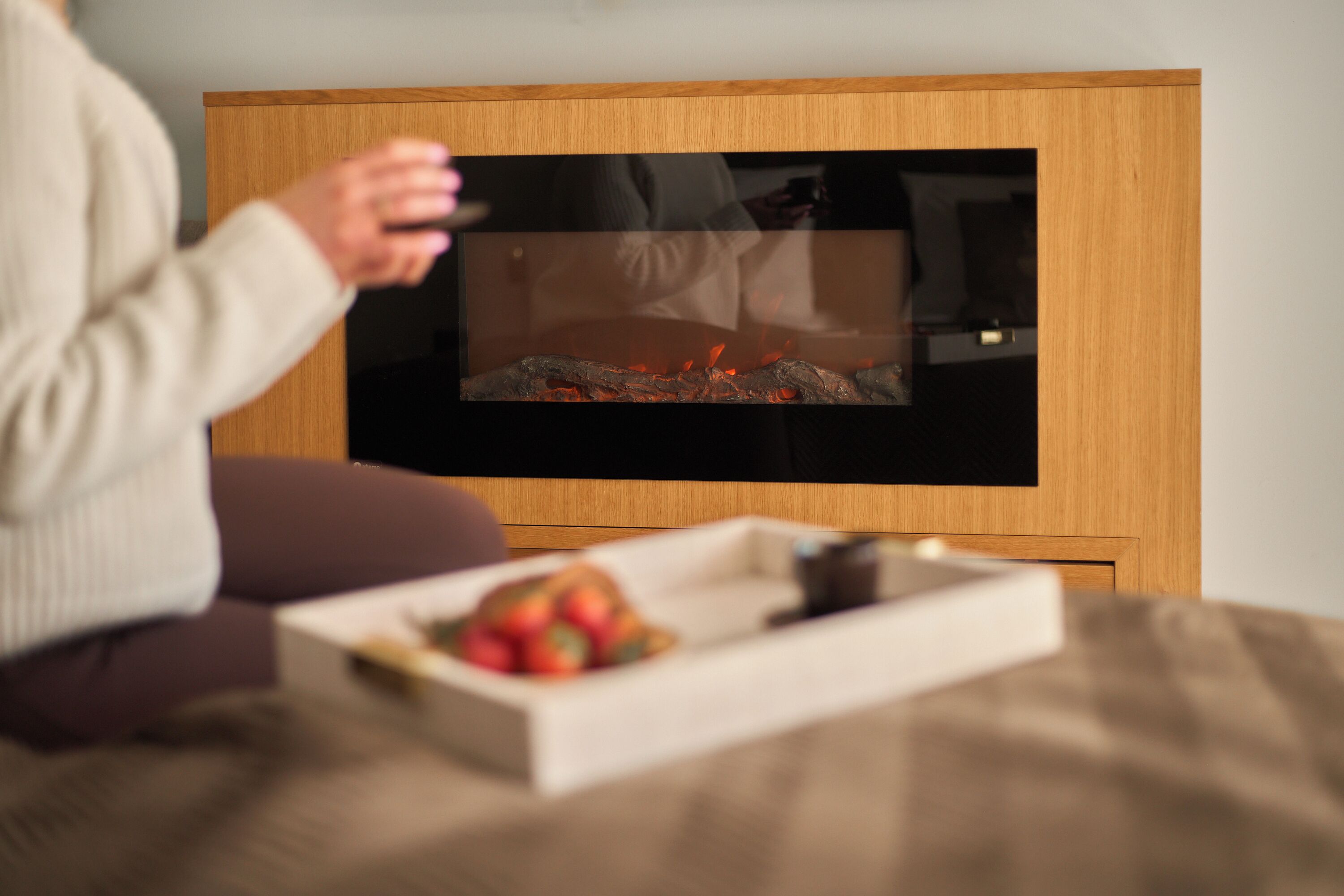 Une personne est assise sur un canapé, regardant la télévision dans une pièce où se trouvent un meuble en bois et une petite table sur laquelle sont disposés des fruits.