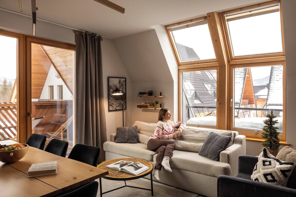 Un salon chaleureux avec un canapé, une table basse et une table à manger avec des chaises. De grandes fenêtres aux cadres en bois offrent une vue sur un paysage enneigé.