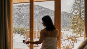 Une personne vêtue d’un peignoir blanc, debout près d’une fenêtre, d’où l’on peut admirer un paysage enneigé et des montagnes.