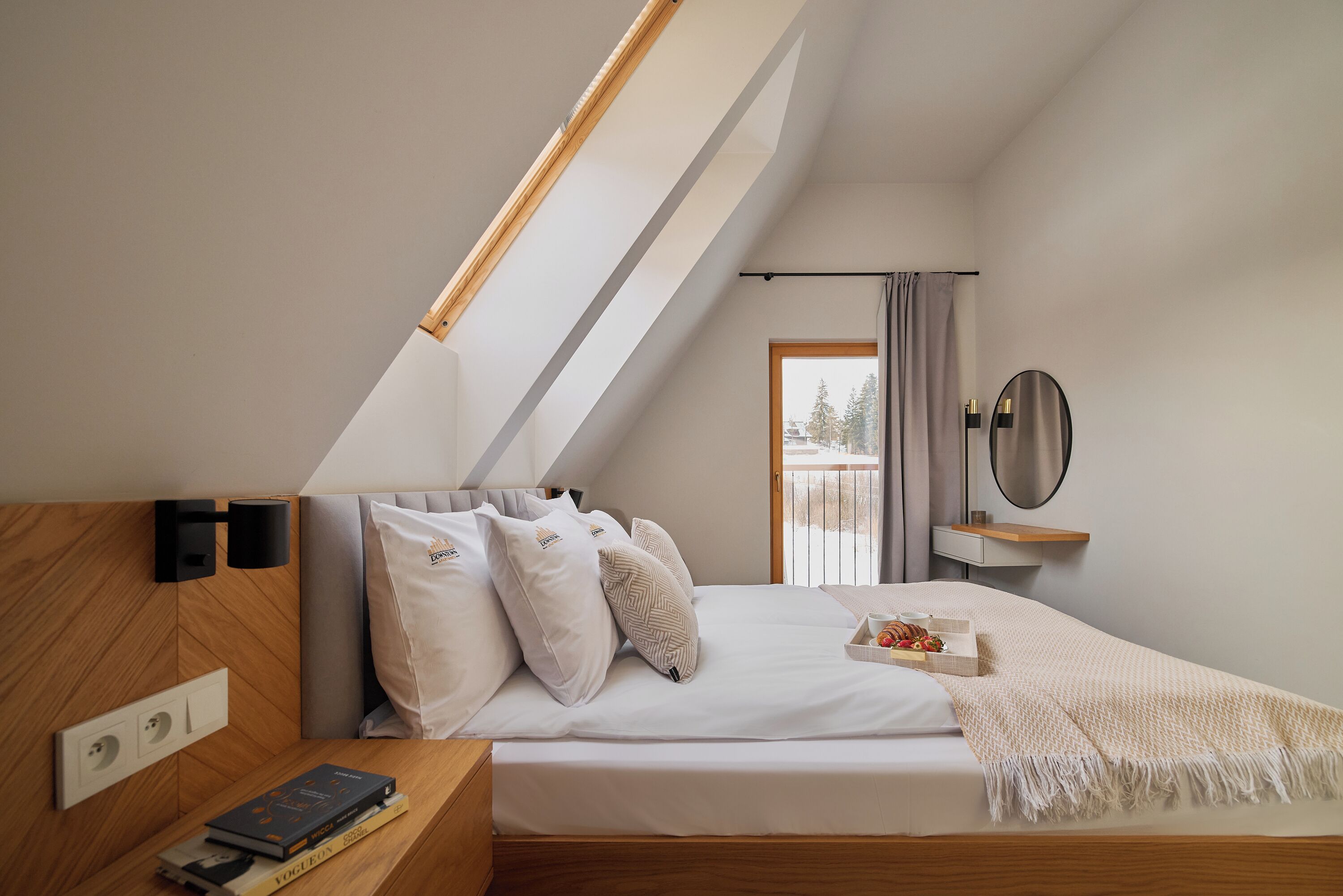 Ein modernes Schlafzimmer mit schrägem Dach, einem Bett mit weißer Bettwäsche, einem Nachttisch mit Büchern, einem Spiegel und einem Fenster mit Vorhängen.