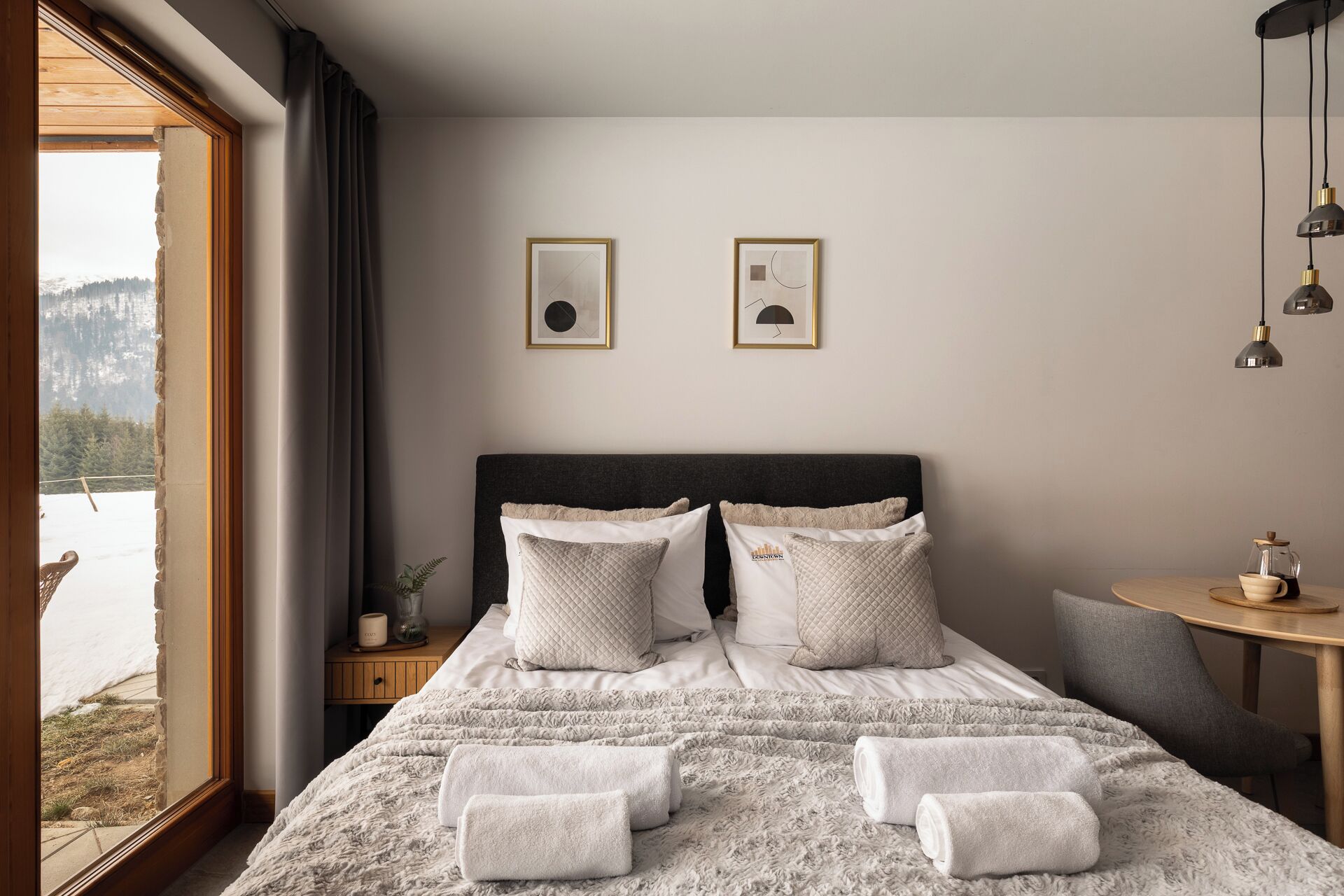 A bedroom with a large bed, two framed pictures on the wall, a bedside table with a lamp, and a view of a snowy landscape through a window.