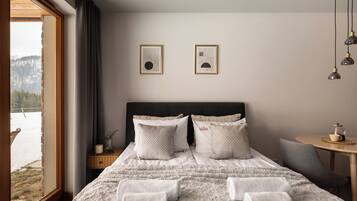 Une chambre avec un grand lit, deux tableaux encadrés au mur, une table de chevet avec une lampe, et une vue sur un paysage enneigé par la fenêtre.