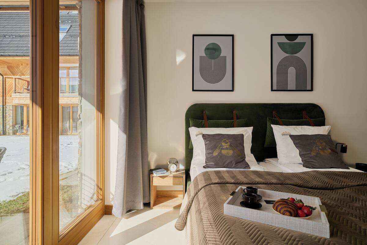 Une chambre avec une grande fenêtre, un lit recouvert d’une courtepointe à rayures, deux oreillers, une table de chevet avec une horloge et des livres, et un plateau avec de la nourriture et des boissons.