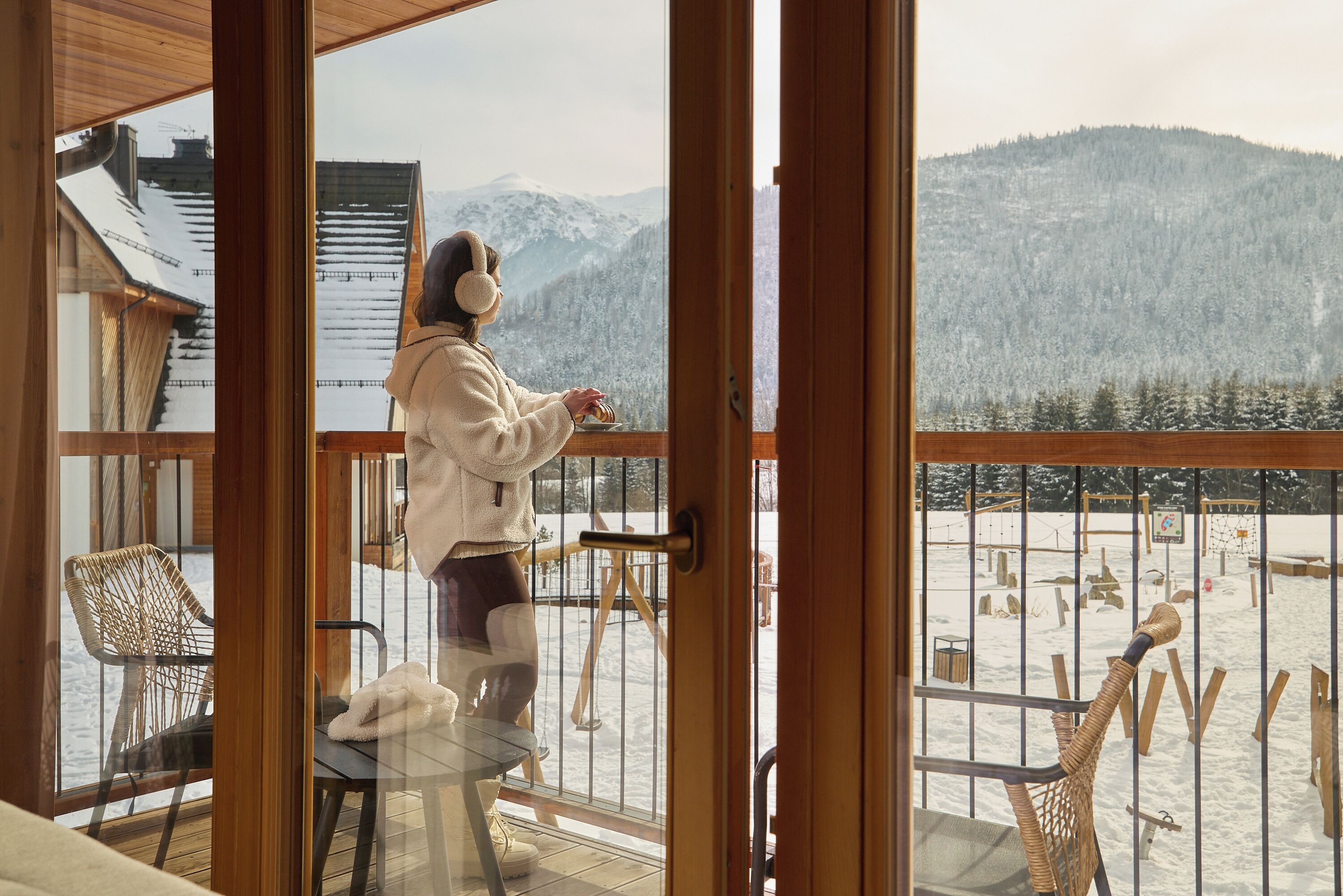 A person standing by a glass door on a balcony overlooking a snowy landscape with mountains.