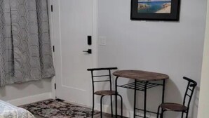 A room with a bed, a small table with two chairs, a door with an 'EXIT' sign, and a framed picture on the wall.