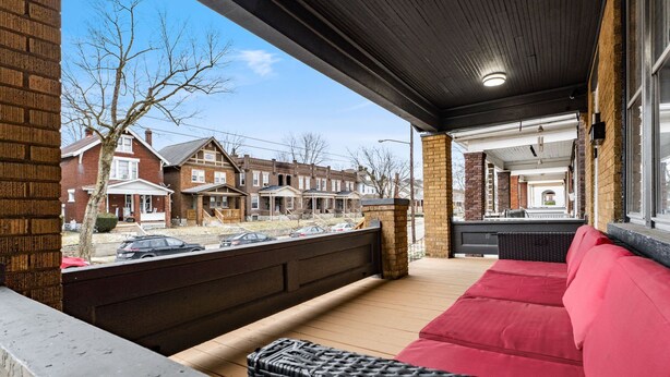 A covered outdoor seating area with a red sofa and wicker furniture, overlooking a row of brick townhouses. - Minutes to Hospital, Cozy 3BR w/ Game Room, Family-Friendly (Columbus)