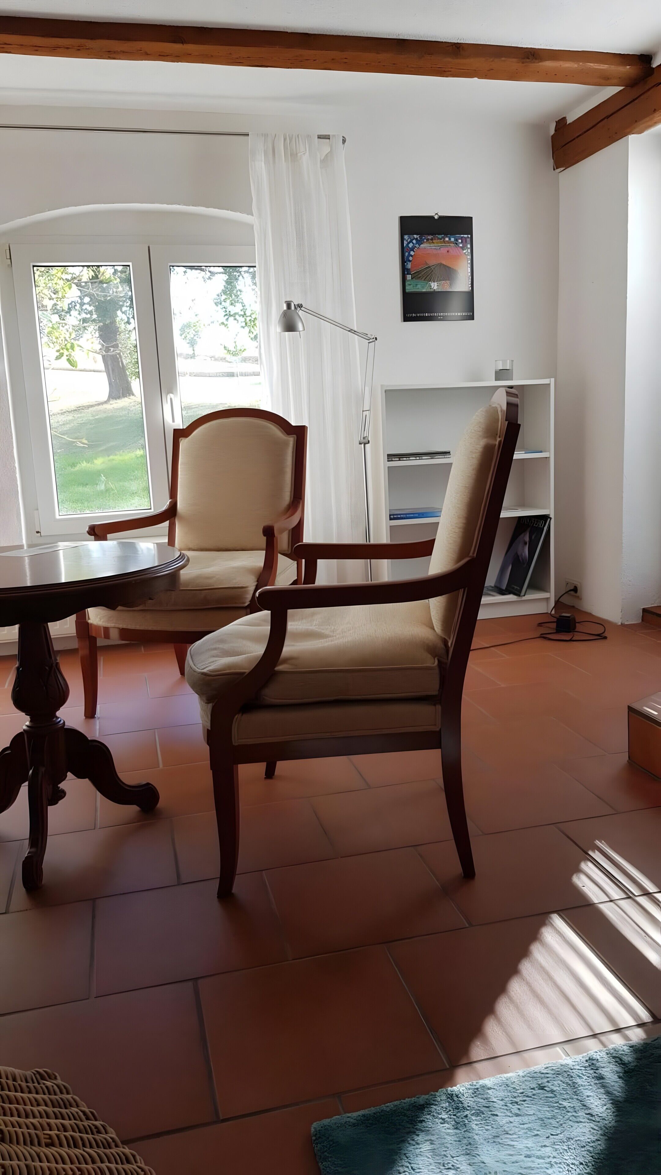 Ein Zimmer mit einem Holzbett, einem Tisch, einer Lampe, einem Bücherregal und einem Fenster mit Vorhängen.