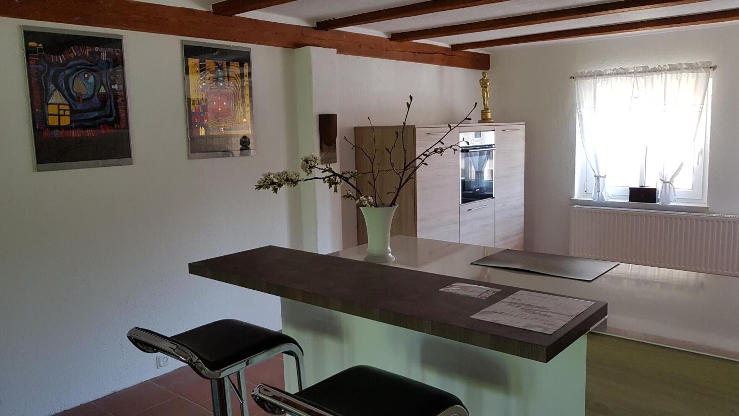 A room with a wooden ceiling, a white cabinet, a vase with branches, a chair, and a window with curtains.