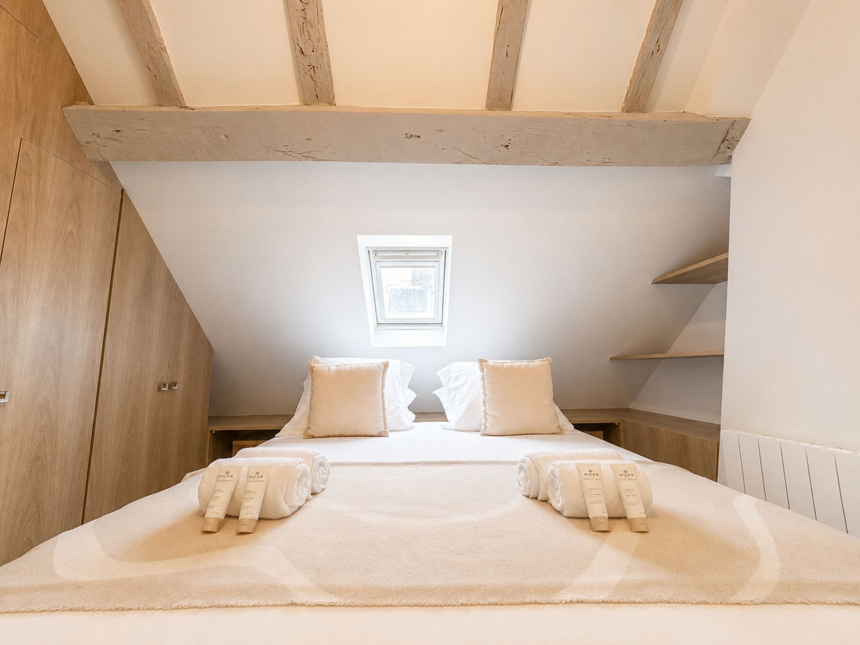 A bedroom with a sloped ceiling, a skylight, a bed with white linens, and a wooden wardrobe.