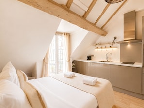 A bedroom with a large bed, a window with curtains, and a kitchenette with a sink and cabinets. - 1531 Bourgeois (Paris)