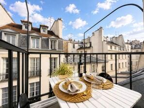 A dining table set with plates, glasses, and a bottle, overlooking a cityscape with buildings and a clear sky. - 1531 Bourgeois (Paris)