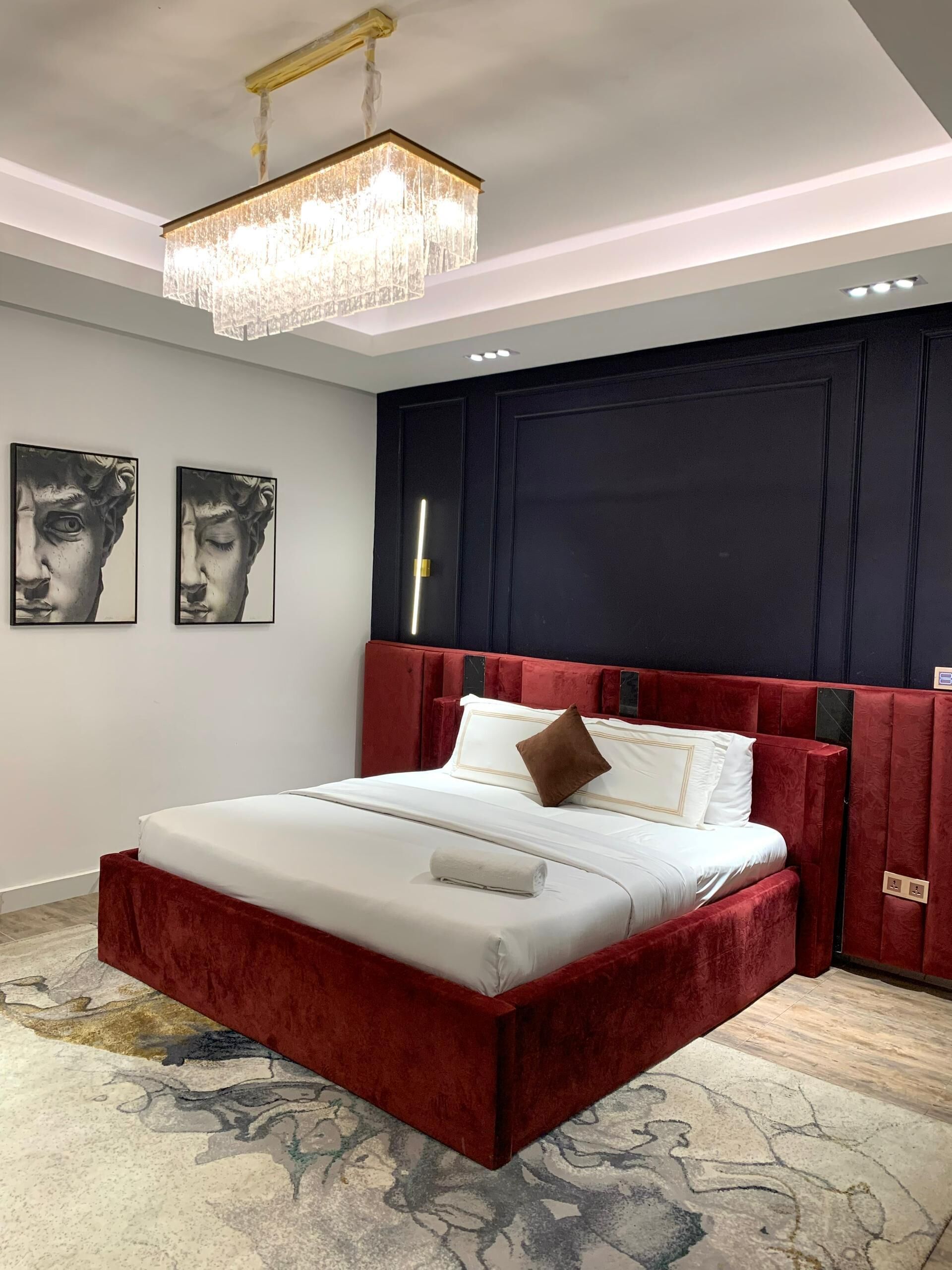 A bedroom with a red velvet bed, a chandelier, and two wall-mounted paintings.