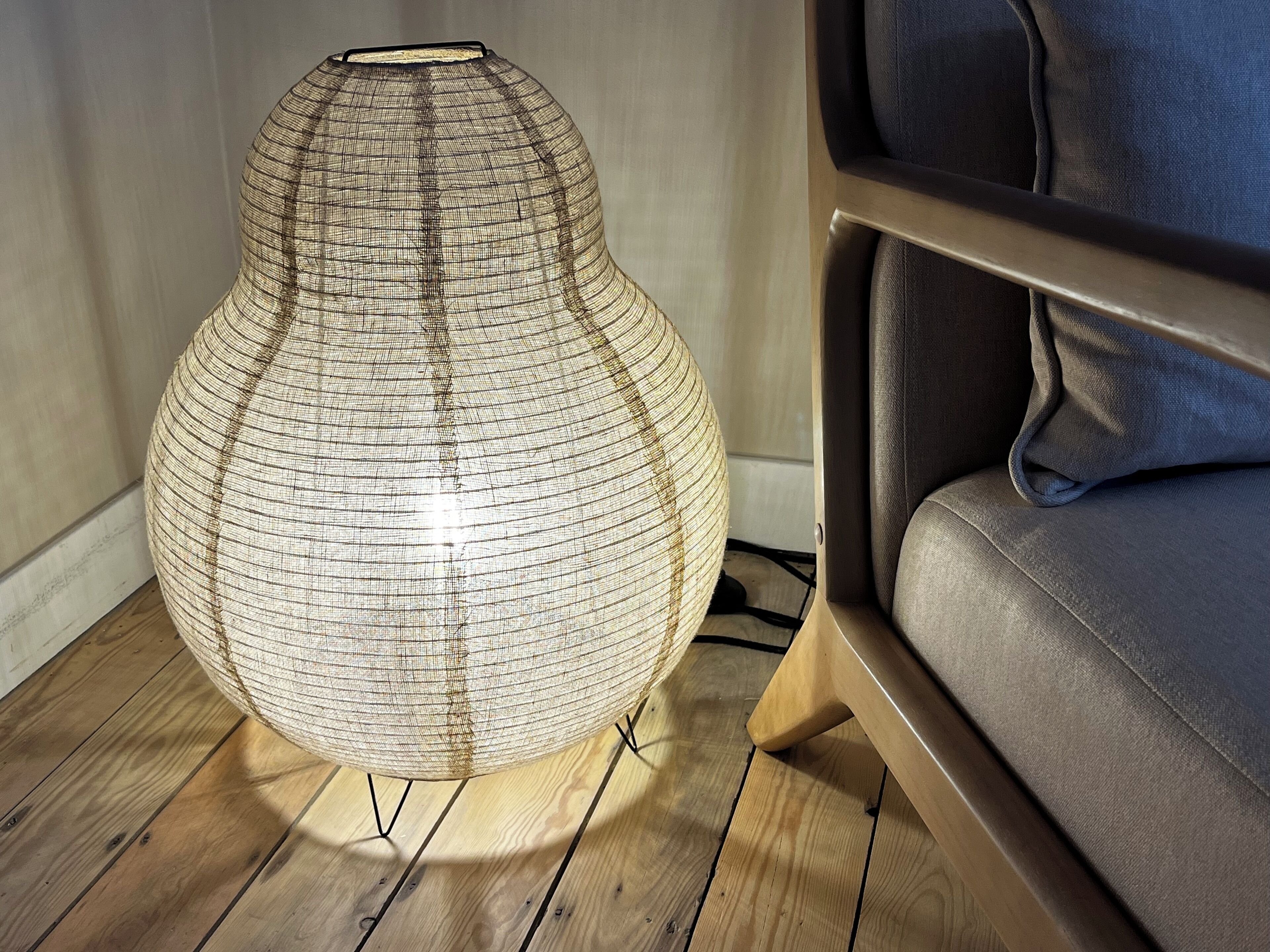 Una lámpara de pie esférica, tejida, con base de madera, ubicada sobre un suelo de madera junto a un sofá gris.