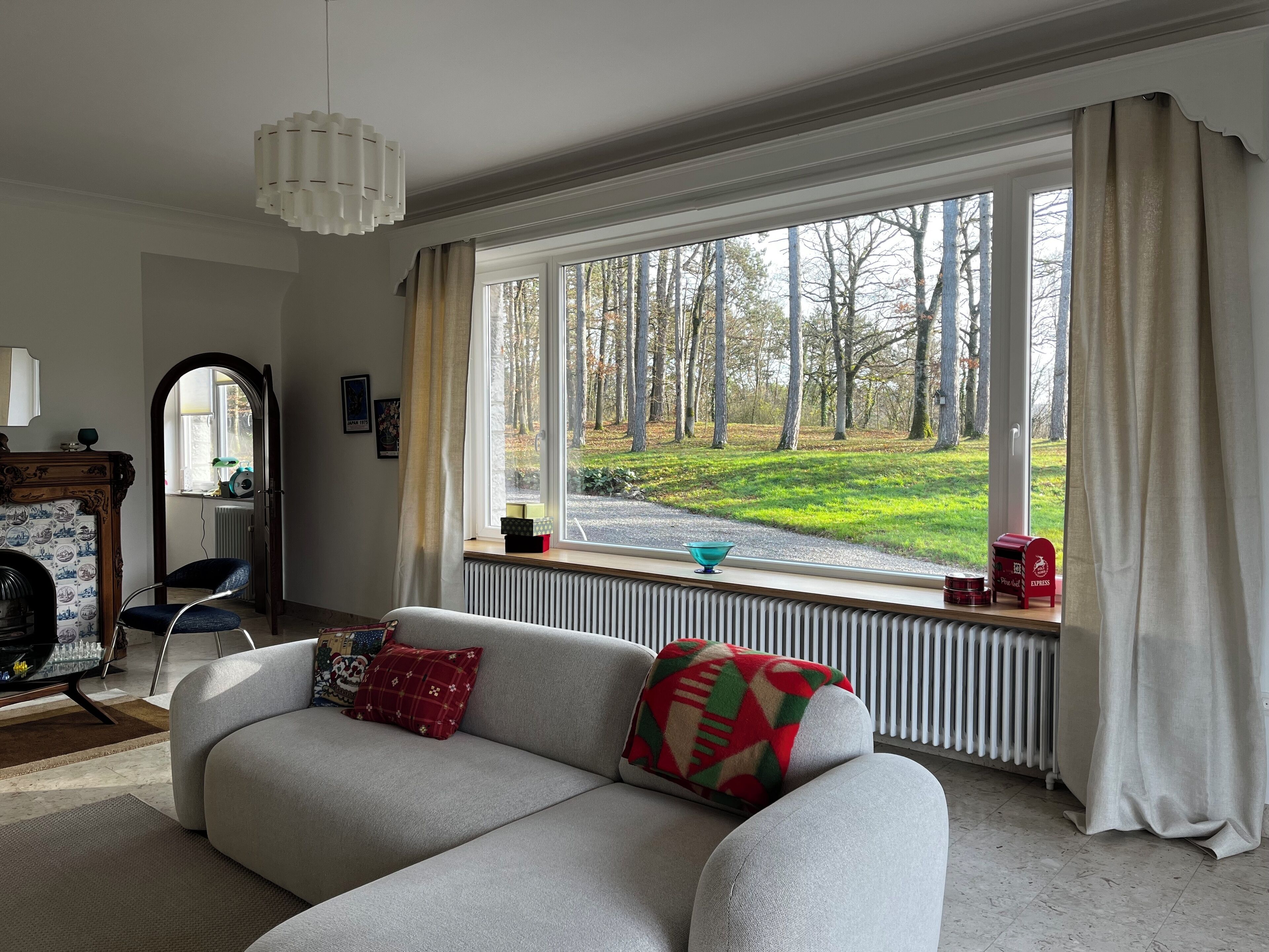 Un salon avec un canapé, un radiateur, une fenêtre donnant sur un parc et une petite table sur laquelle se trouvent un bol et une boîte rouge.