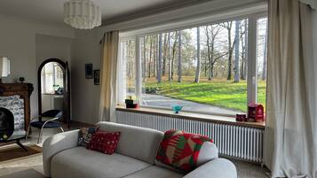 Un salon avec un canapé, un radiateur, une fenêtre donnant sur un parc et une petite table sur laquelle se trouvent un bol et une boîte rouge.