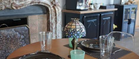 Een eettafel gedekt met een bord, twee glazen en een glas blauwe vloeistof. Op de tafel staat een mozaïeklamp, en een koffieapparaat staat op de achtergrond.