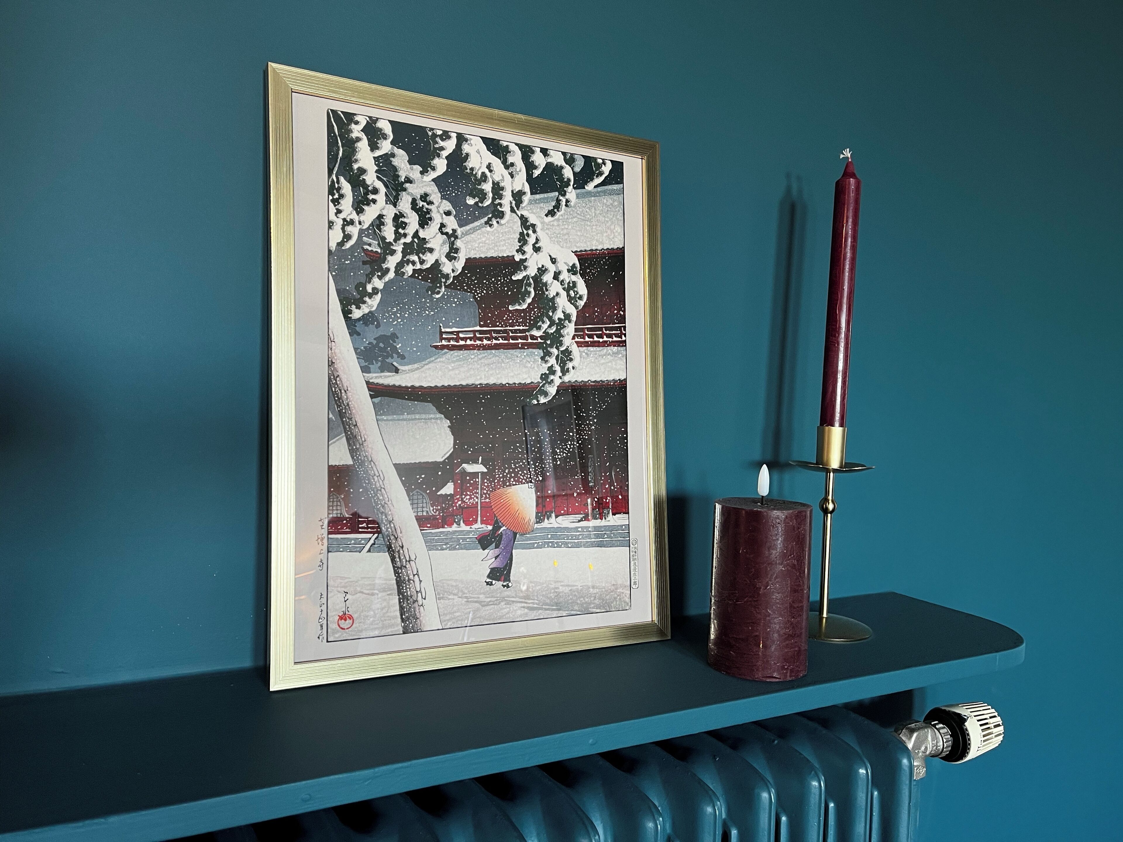 A framed artwork of a snowy scene with a person in traditional attire, a red candle, and a radiator below.
