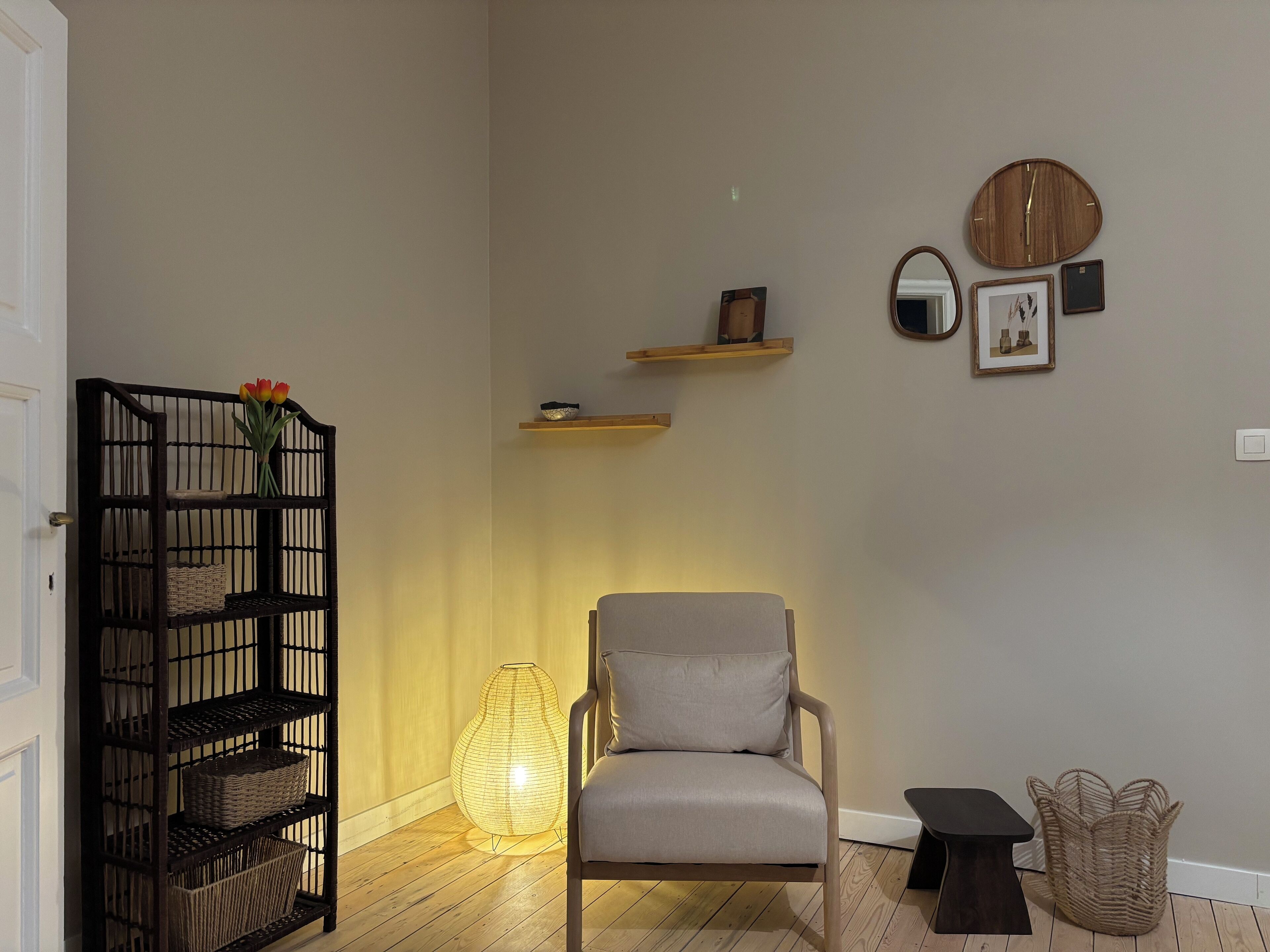 Una habitación con piso de madera, un sillón gris, un estante negro con cestas, una lámpara de pie tejida y estantes en la pared con objetos decorativos.