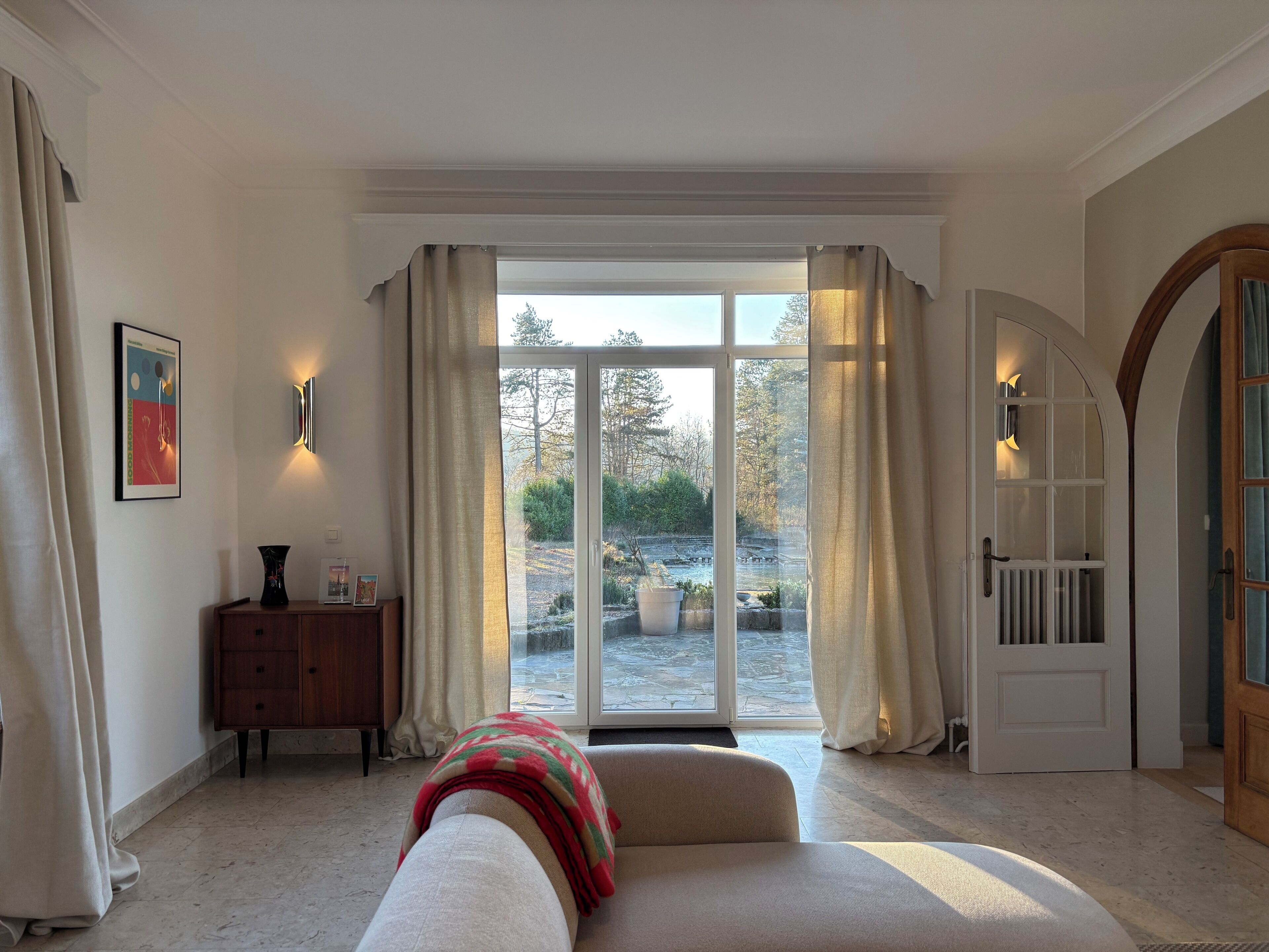 Une pièce dotée d’une grande porte-fenêtre coulissante donnant sur un jardin, d’un canapé avec un plaid, d’une armoire en bois et d’un tableau encadré accroché au mur.