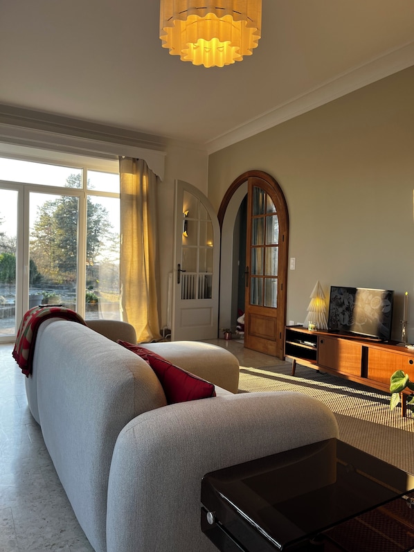 A modern living room with a large sofa, a coffee table, and a television on a wooden stand. There is a large window with curtains, a door with an arched design, and a chandelier. - Aux étangs fleuris (Rochefort)