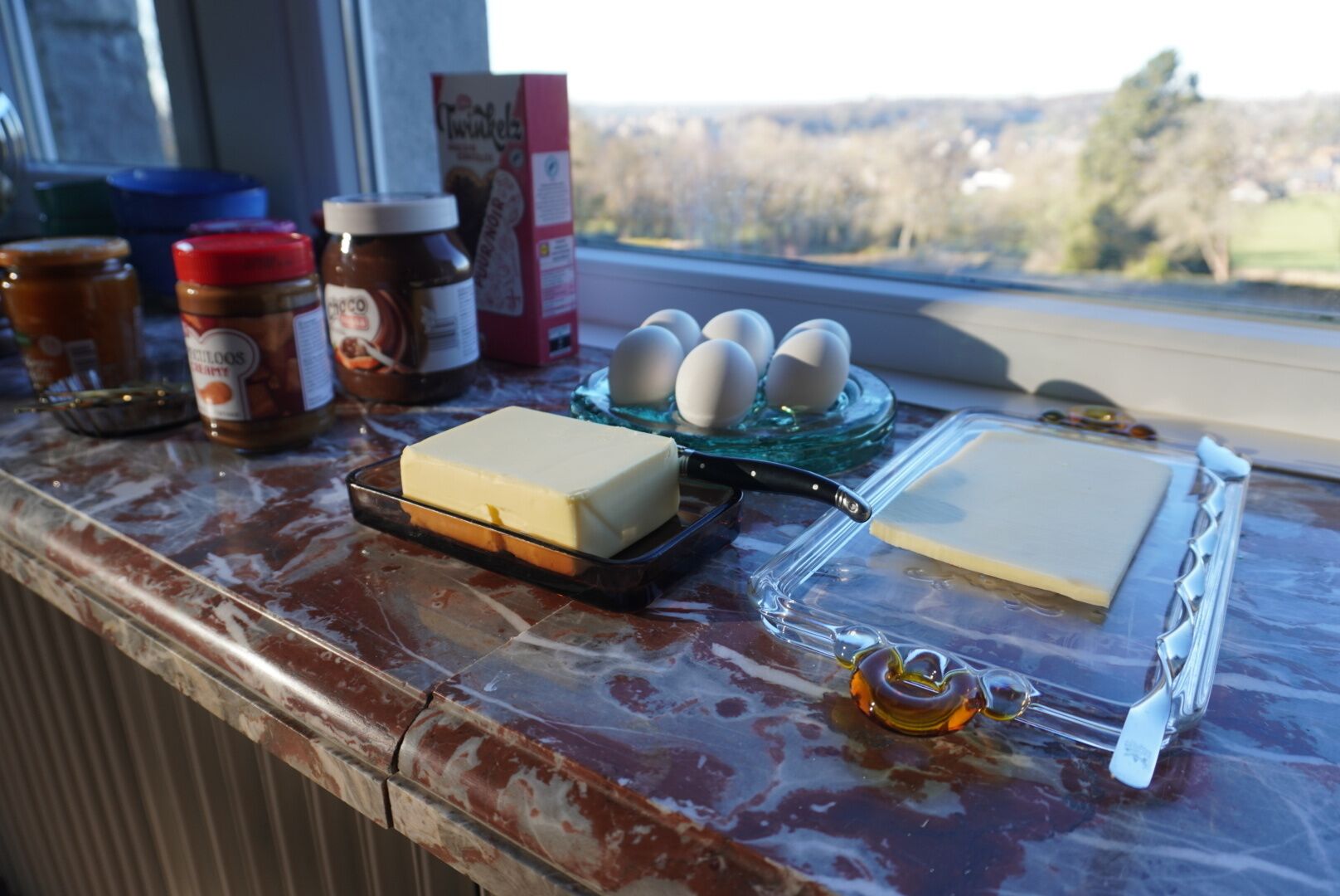 Un plan de travail de cuisine avec un morceau de beurre, des œufs, des pots de confiture et une boîte de céréales.