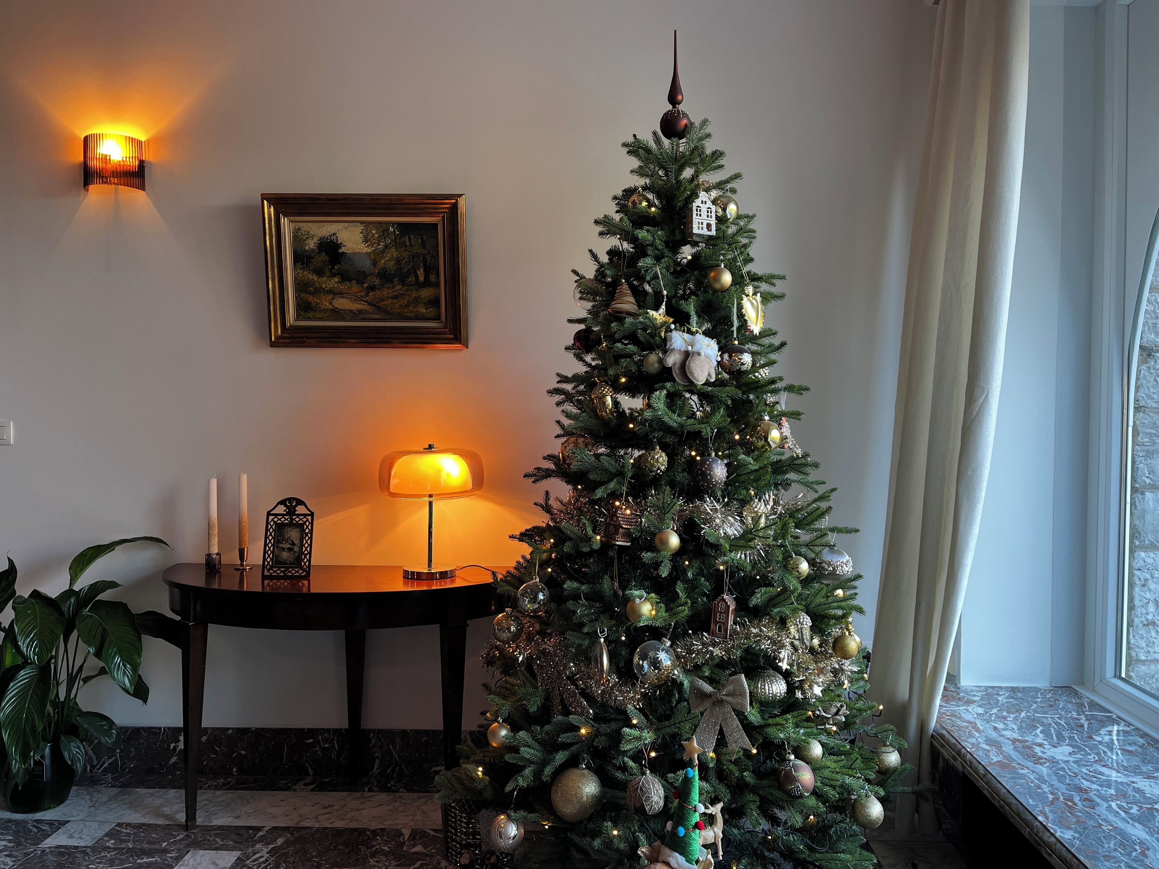 Een kerstboom met versieringen, een lamp, een lijst met een foto en een potplant in een kamer met een marmeren vloer.