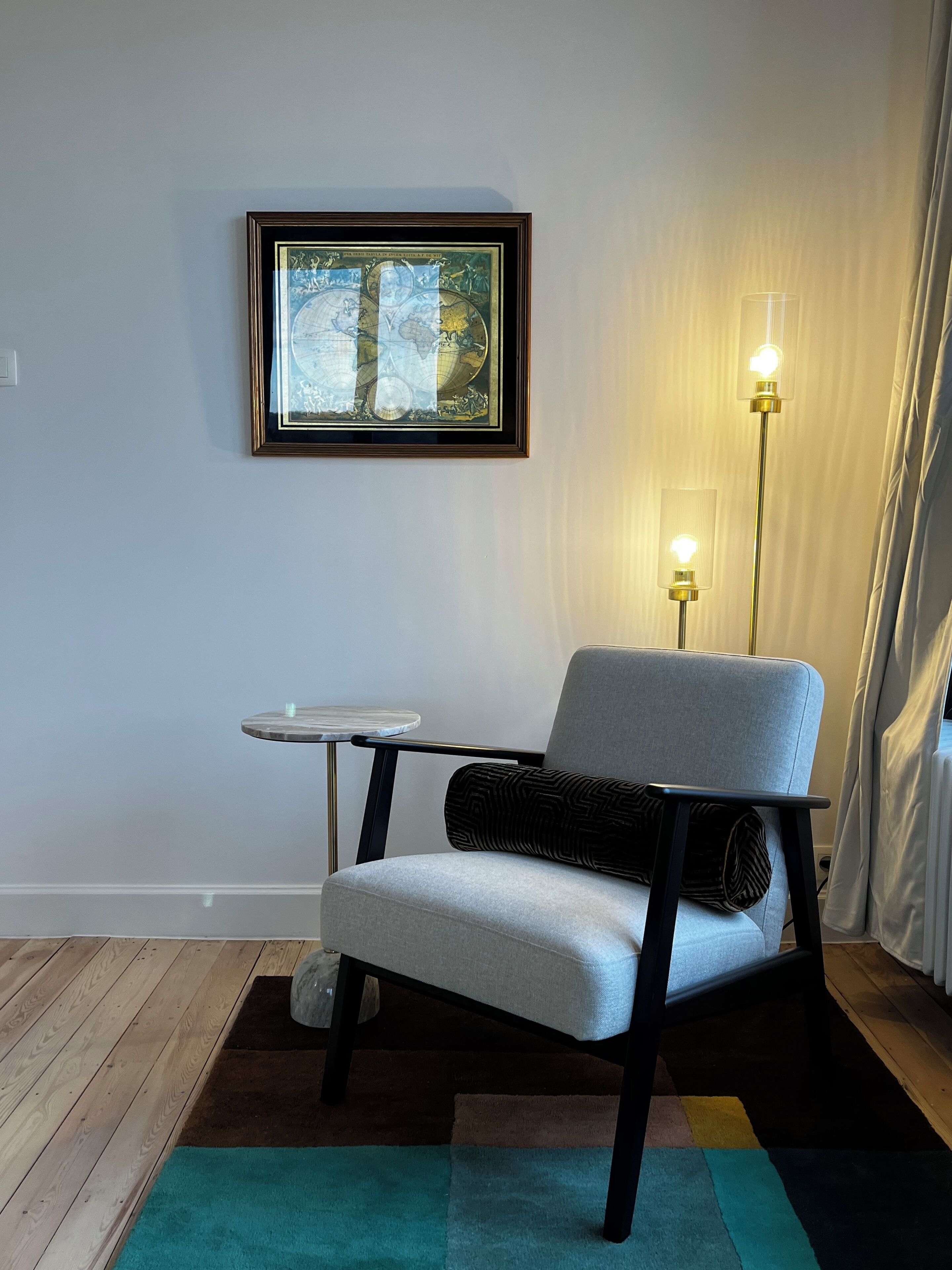 A modern living room with a grey armchair, a small round table, a framed map on the wall, and two floor lamps.
