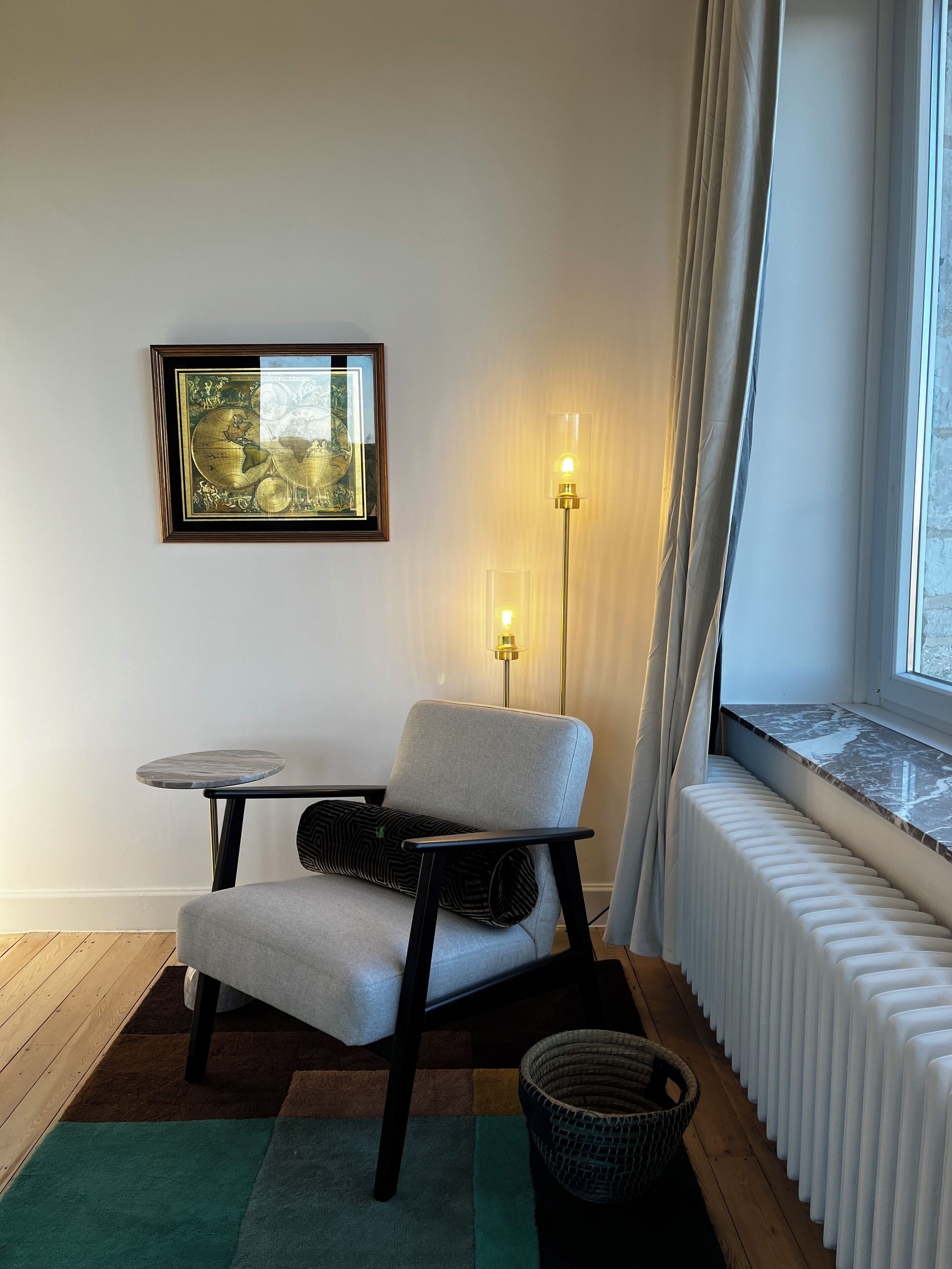 Una habitación moderna con un sillón gris, una mesita redonda pequeña, un mapa enmarcado en la pared, una lámpara de pie y un radiador.