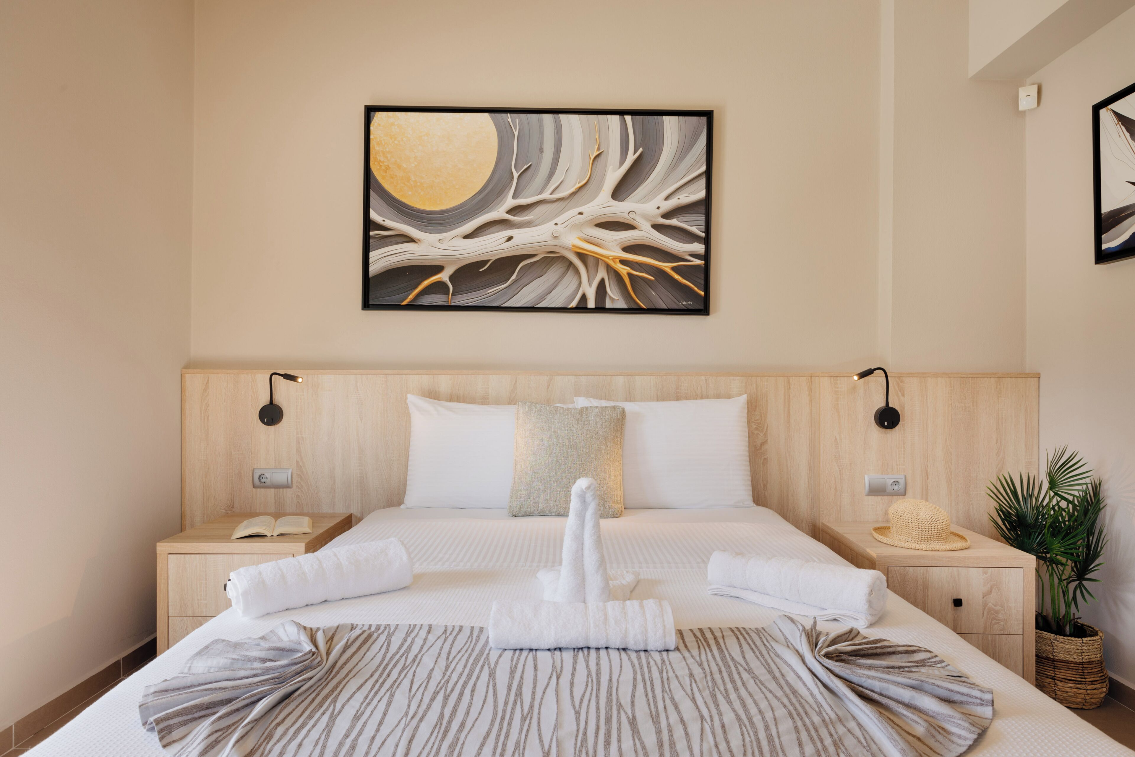 Una habitación de hotel con una cama grande, una mesita de noche, una obra de arte colgada en la pared, una lámpara de noche, una planta y una cesta tejida.