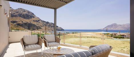 A balcony with wicker furniture, a table, and a view of the sea and mountains.