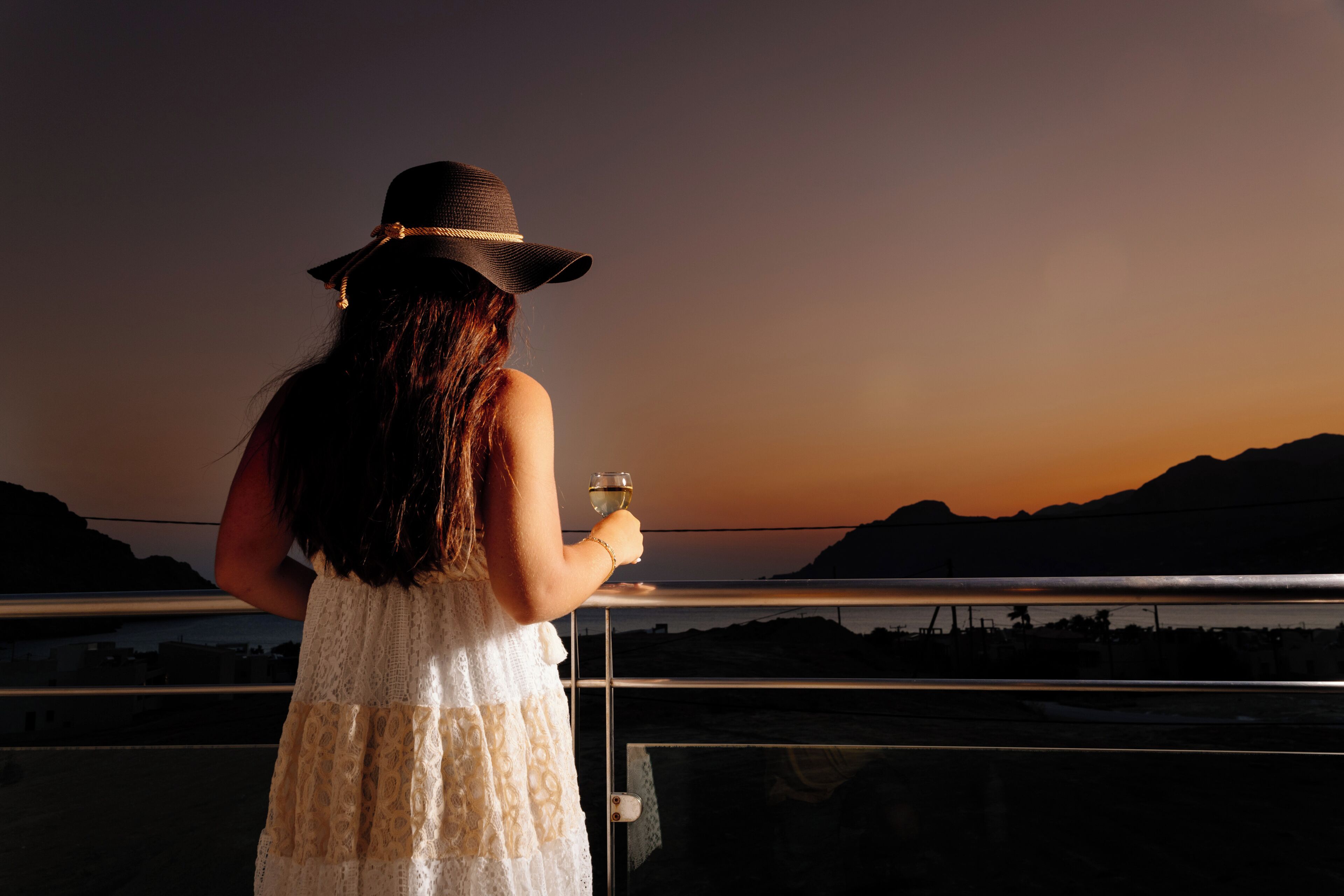 Una persona con vestido y sombrero blancos sostiene un vaso, con vistas a un paisaje con montañas y una masa de agua.