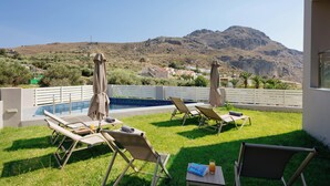 Zona de piscina con sillones de playa y sombrillas, una mesita con un libro y un vaso de jugo, y una vista de montañas y edificios al fondo.
