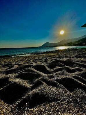 A beach with the sun setting over the ocean, casting shadows on the sand. - Lamon (Agios Vasileios)