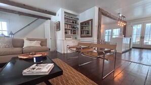 Amplia sala de estar con sofá, mesa de centro y mesa de comedor con sillas. Se ve una escalera que conduce a otro nivel, una estantería y una cocina visible a través de una puerta abierta.