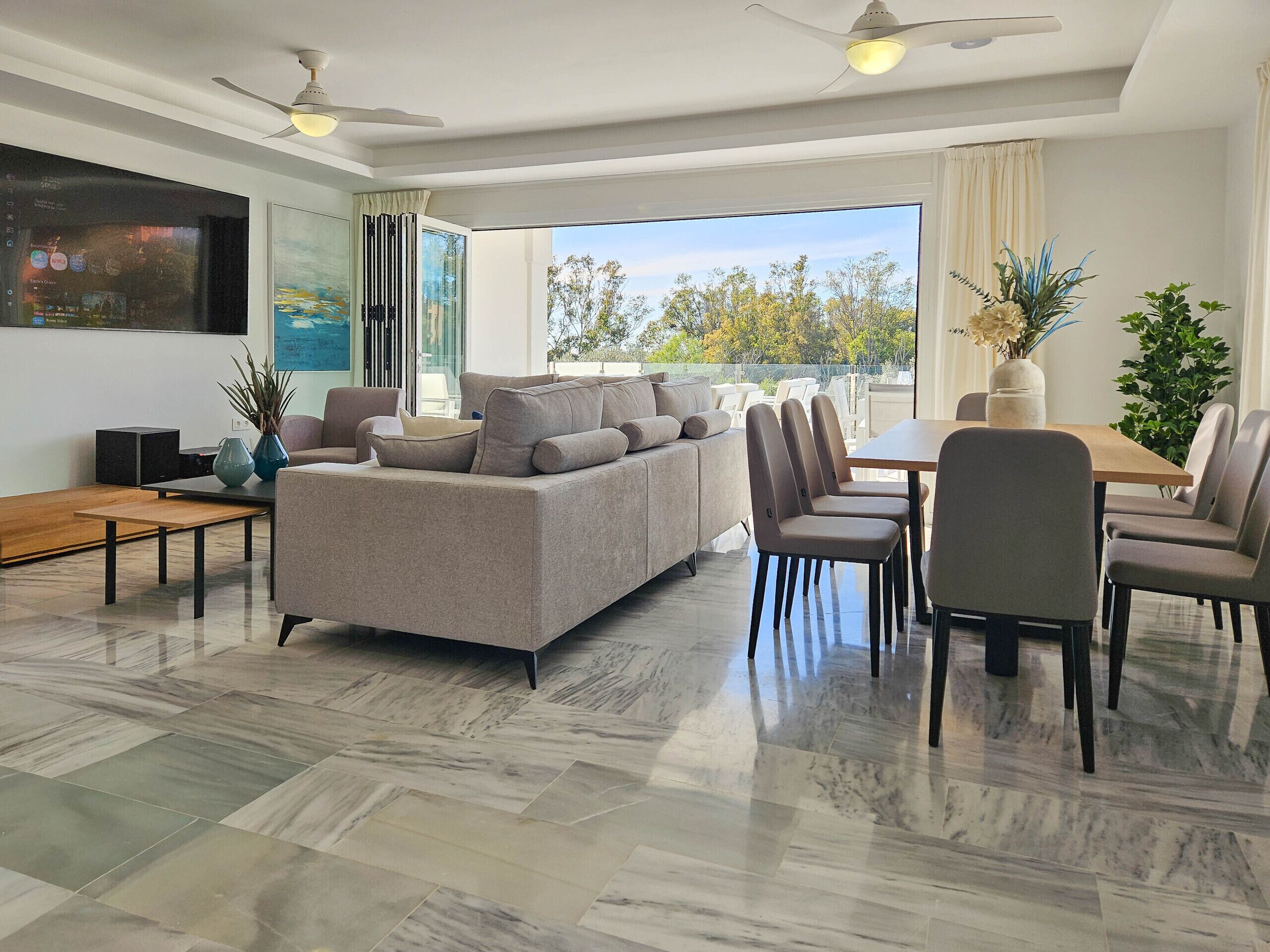 Ein modernes Wohnzimmer mit einem Ecksofa, einem Esstisch und Stühlen, großen Fenstern mit Blick auf Bäume und einem Deckenventilator.